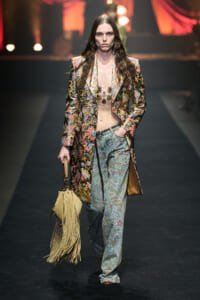 Model walking the runway in a ornate floral-patterned suit with a long jacket, matching patterned pants, and a fringe tan bag.