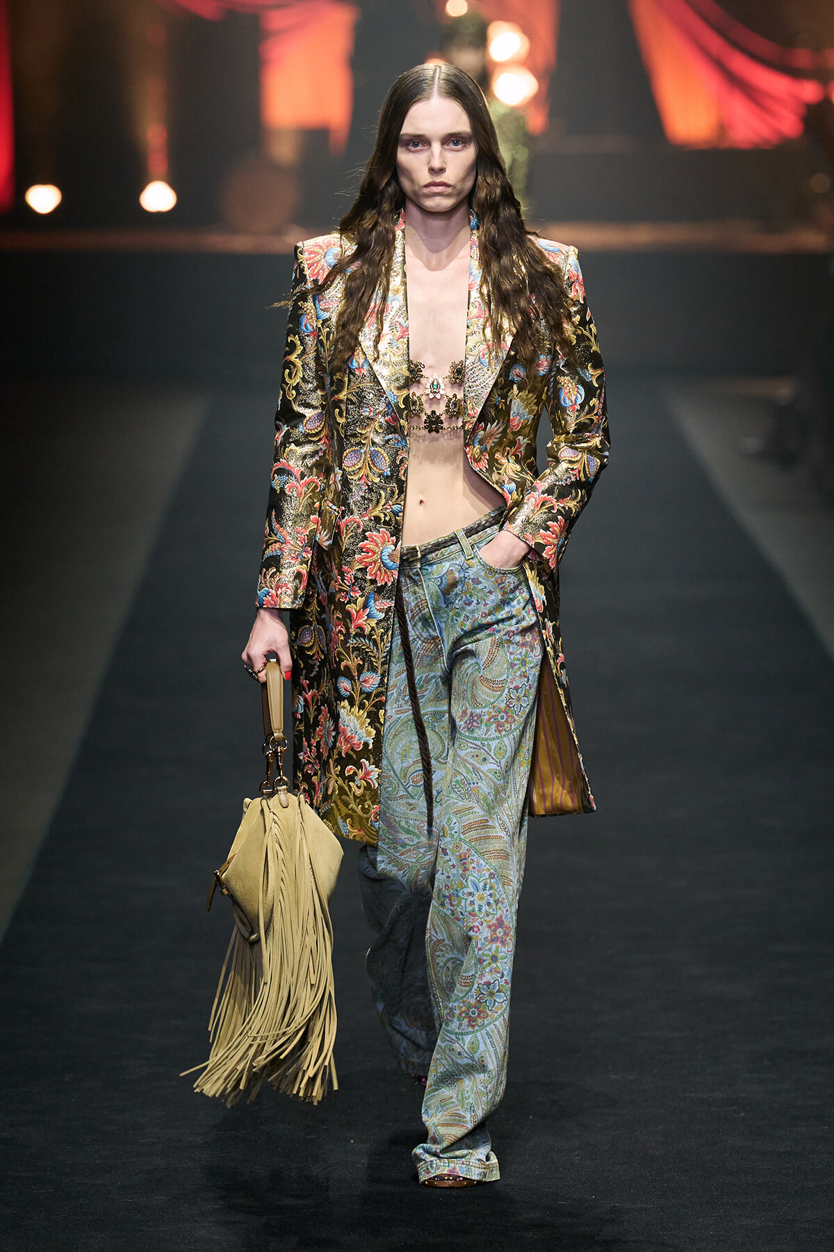 Model walking the runway in a ornate floral-patterned suit with a long jacket, matching patterned pants, and a fringe tan bag.