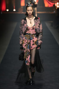 Model walks a fashion runway wearing a pink floral dress with a sheer black overlay and mismatched accessories, carrying a black beaded bag.