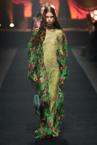 Model walking a runway in a long green paisley dress with layered sheer fabric and a small blue purse.