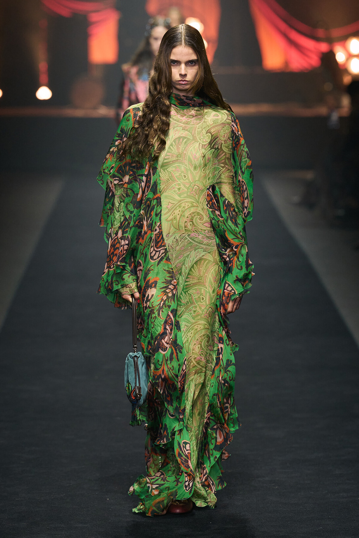 Model walking a runway in a long green paisley dress with layered sheer fabric and a small blue purse.