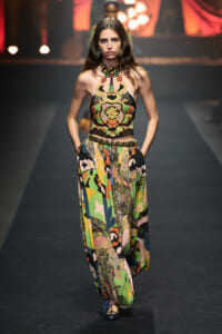 Model walks the runway wearing a vibrant embroidered halter top and wide, colorful patterned pants with a metallic belt.