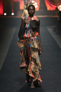 Model walks the runway in a colorful, layered bohemian dress with black sheer top and vibrant scarf-like skirt, accessorized with a chunky multicolor scarf necklace and small bag.