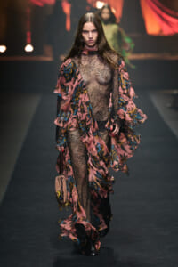 Model walking a fashion runway in a pink floral lace gown with a sheer bodice and flowing layered sleeves, paired with black tights and a small beige handbag.