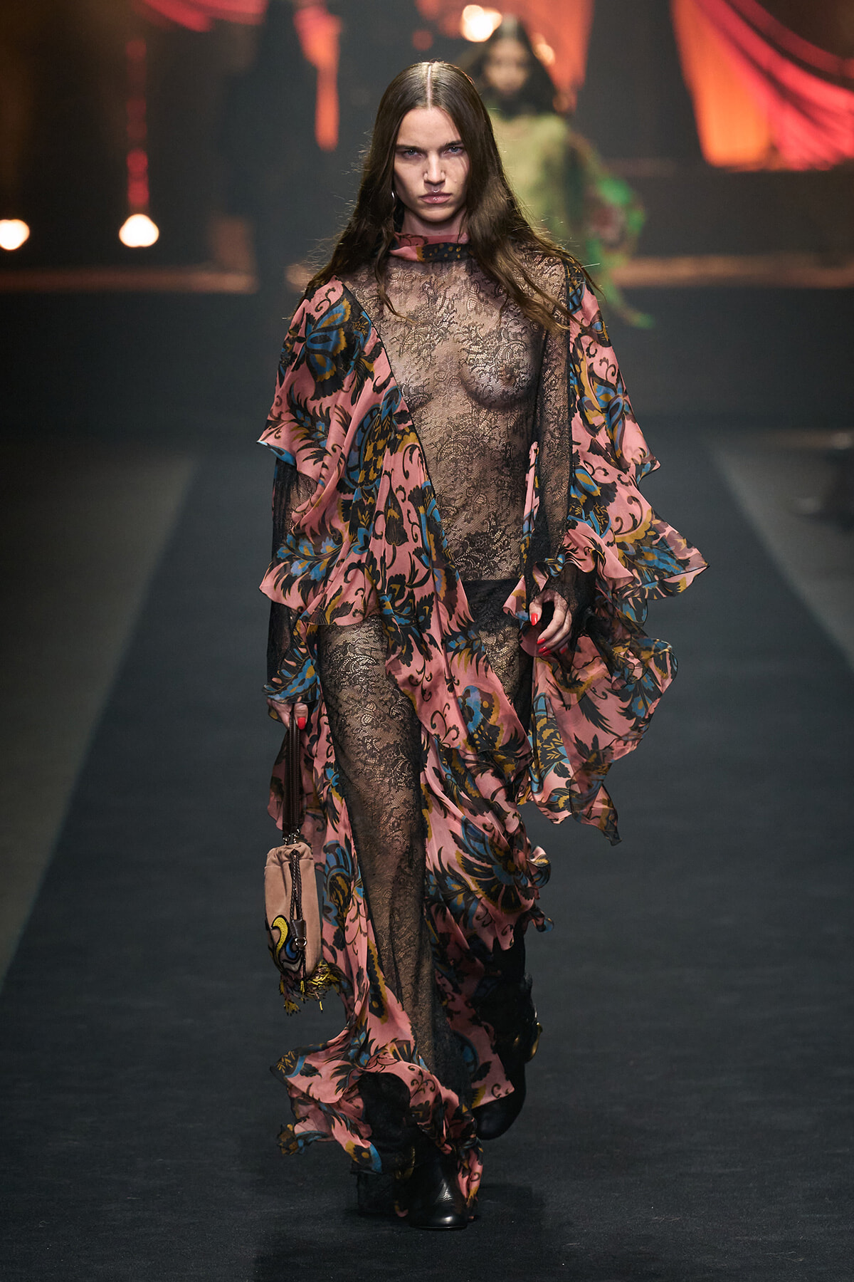 Model walking a fashion runway in a pink floral lace gown with a sheer bodice and flowing layered sleeves, paired with black tights and a small beige handbag.
