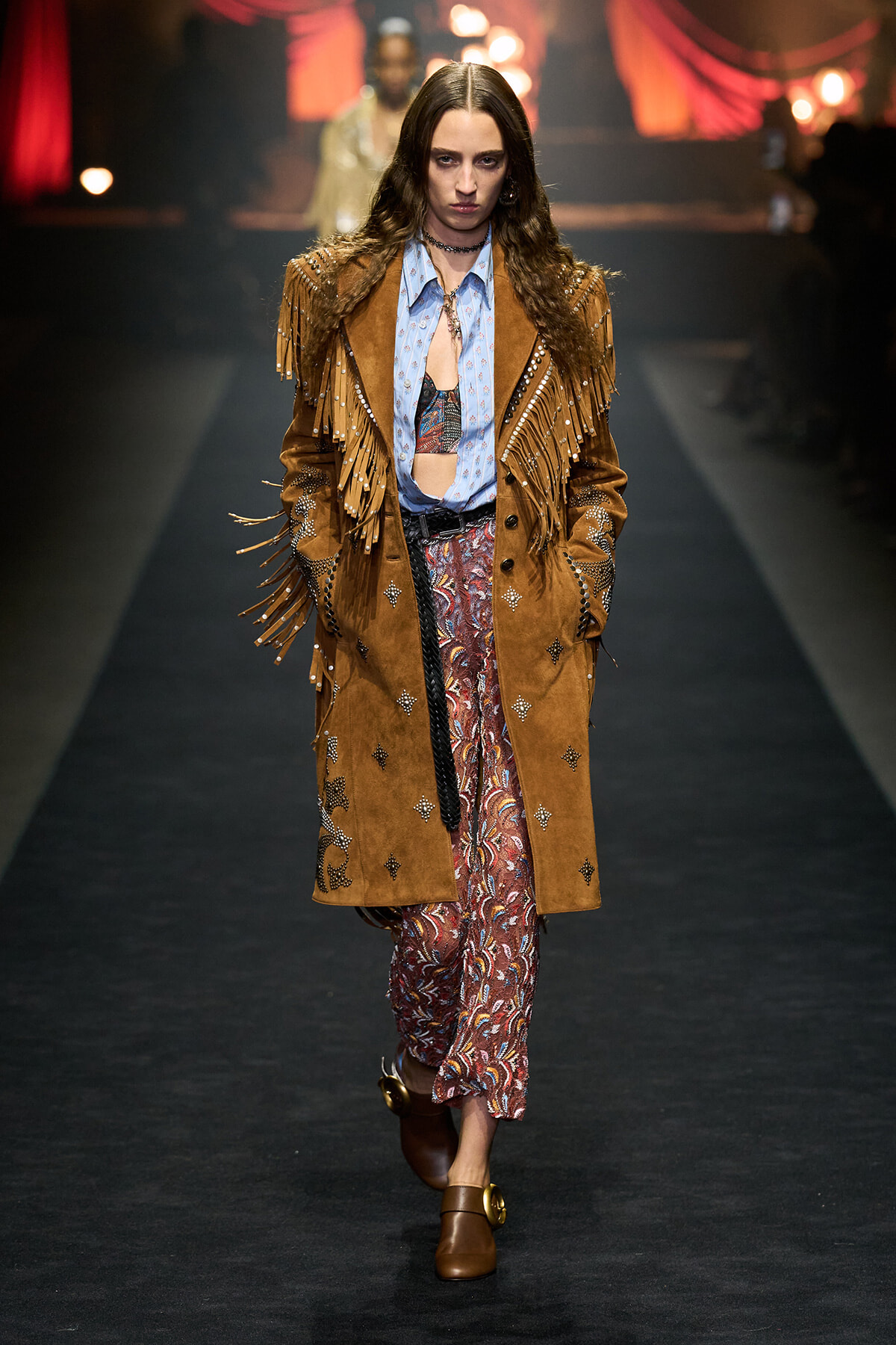 Model walks the runway in a brown suede fringe coat with bead accents over a blue patterned blouse and paisley pants.