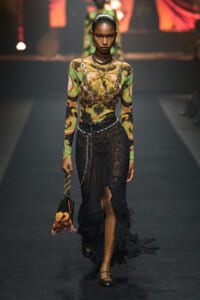 Runway model wearing a colorful swirl-patterned top and black crochet skirt with fringe, layered jewelry, and a small embroidered bag.