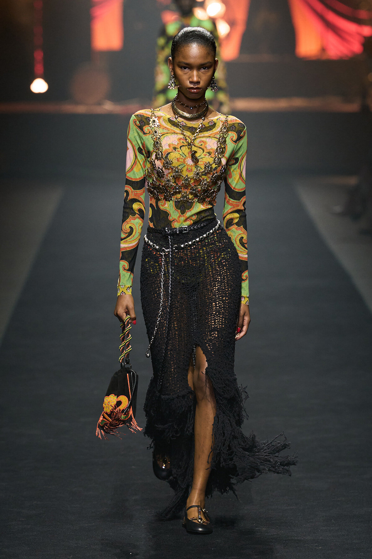 Runway model wearing a colorful swirl-patterned top and black crochet skirt with fringe, layered jewelry, and a small embroidered bag.