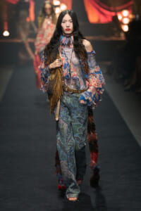 Runway model in boho outfit with sheer floral top, paisley wide-leg pants, brown fringe handbag, and layered accessories.