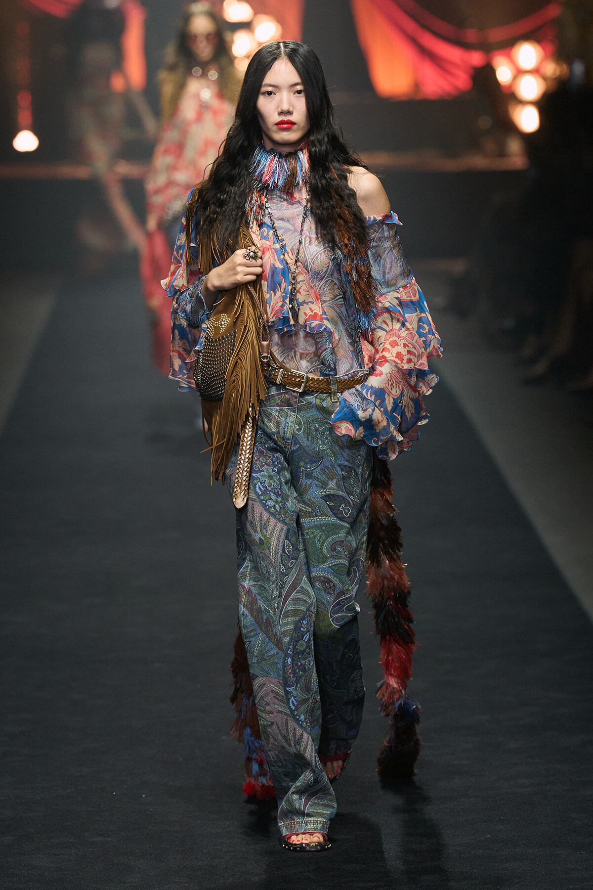 Runway model in boho outfit with sheer floral top, paisley wide-leg pants, brown fringe handbag, and layered accessories.