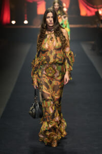 Runway model walking a catwalk in a sheer, multicolored paisley dress with ruffled layers, carrying a black handbag.