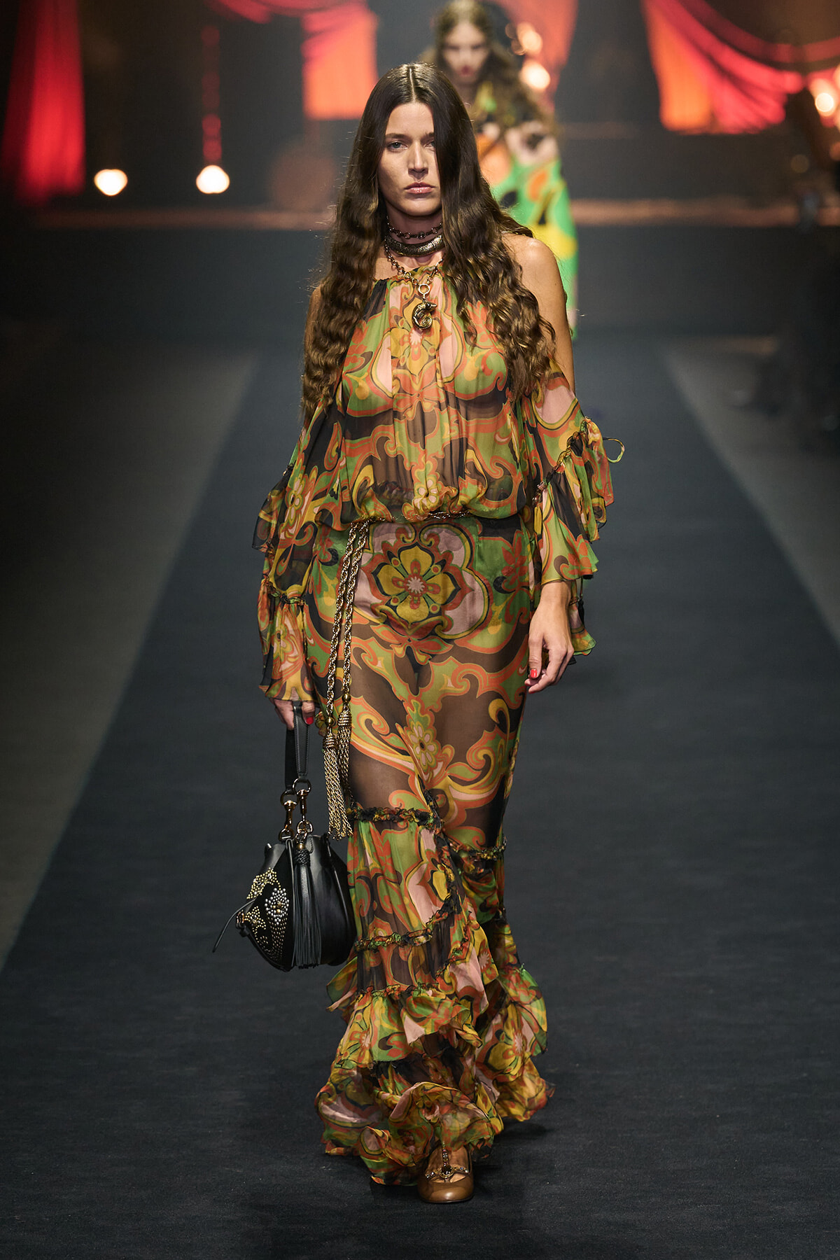 Runway model walking a catwalk in a sheer, multicolored paisley dress with ruffled layers, carrying a black handbag.