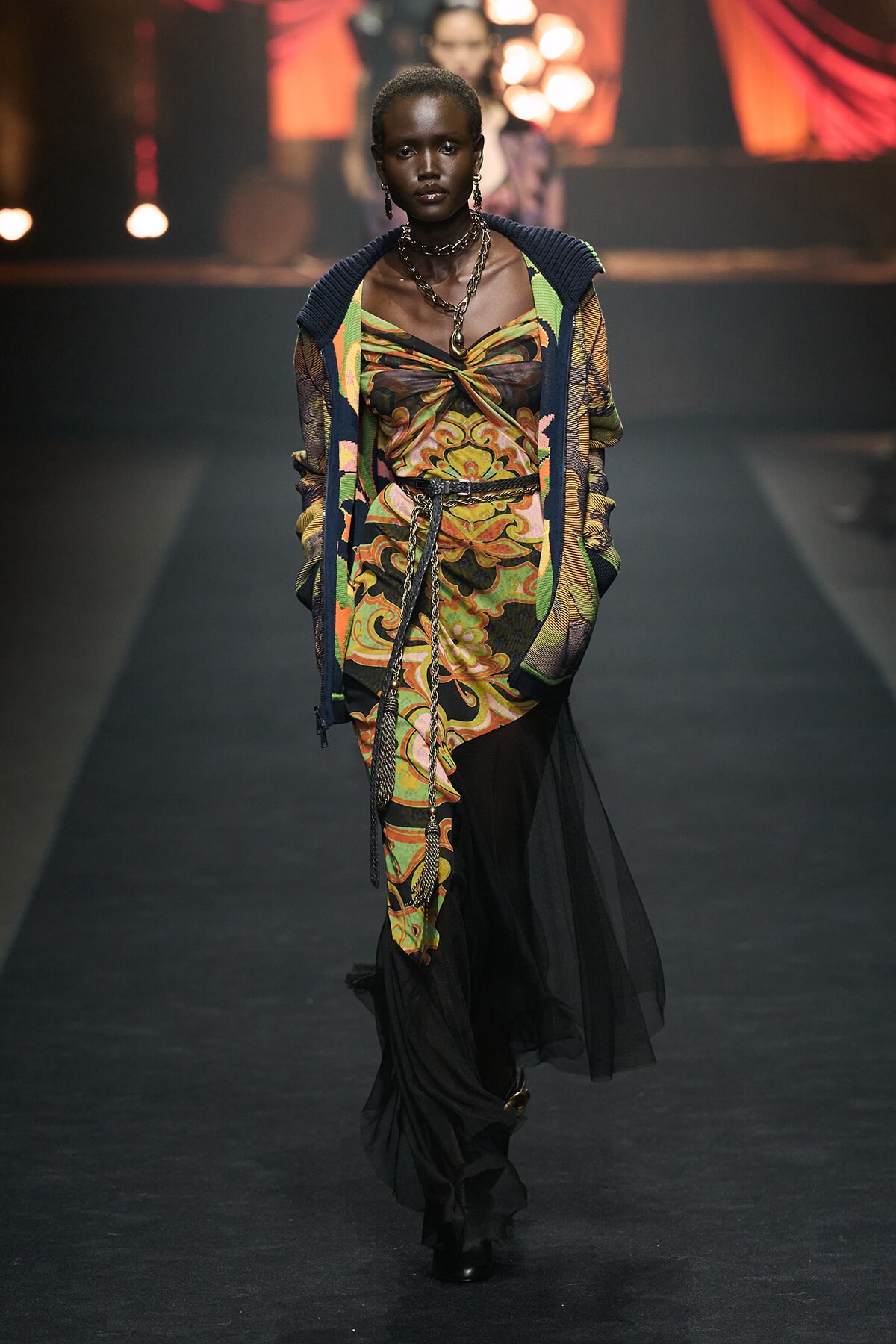 Model walks the runway in a vibrant swirling‑print dress with a sheer black overlay and a knit cardigan.