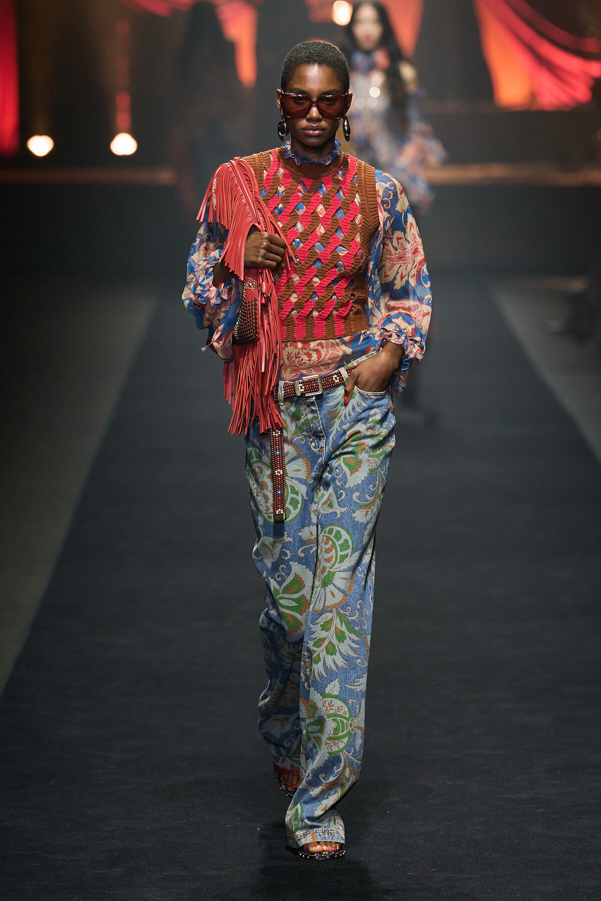 Model walks the runway in a pink fringe bag, pink crochet vest over a blue paisley blouse, and matching wide-leg pants with accessories.
