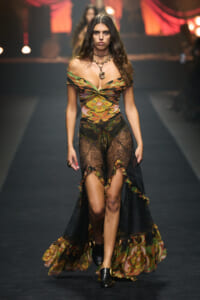 Model walks the runway in a bohemian-patterned off-shoulder gown with black lace overlay and layered necklaces.