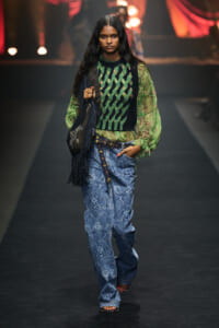 Model walks a runway wearing a green paisley sheer blouse under a black-green knit vest, blue patterned denim jeans, and a studded belt with a fringe bag over the shoulder.