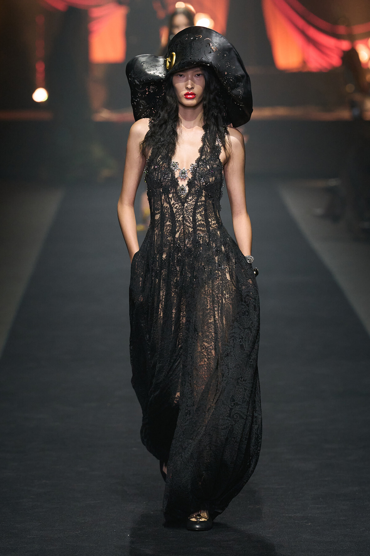 Model on a fashion show runway wearing a sheer black lace gown and an oversized black hat.