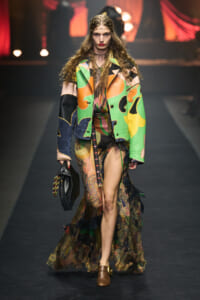 Model walks a runway in a bold, colorful patchwork coat over a sheer patterned gown with a high slit. She carries a black fringed bag and wears brown shoes.