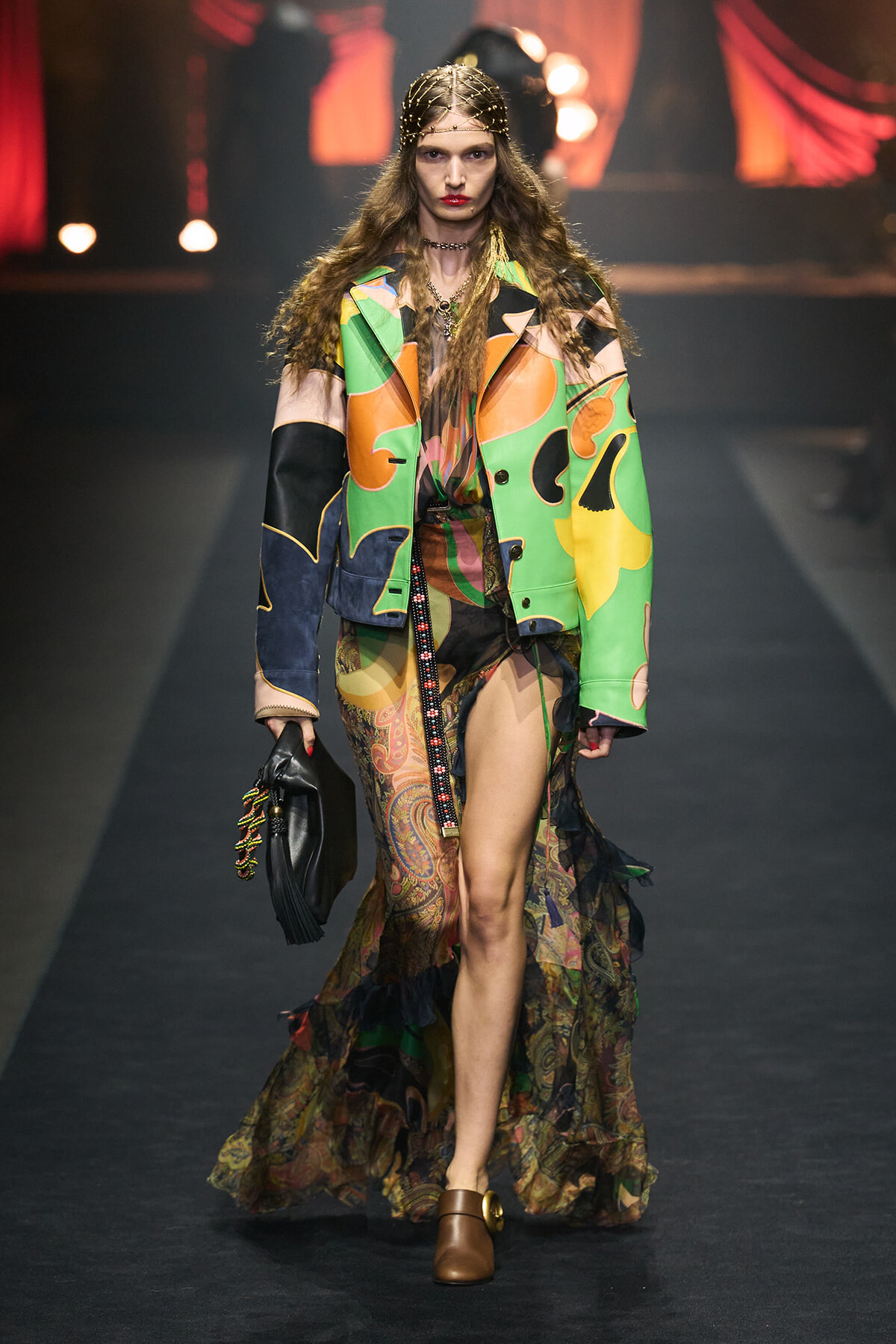 Model walks a runway in a bold, colorful patchwork coat over a sheer patterned gown with a high slit. She carries a black fringed bag and wears brown shoes.