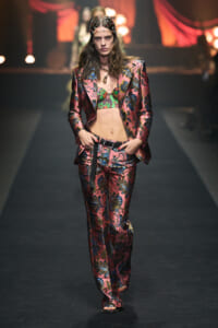 Model walking a fashion runway in a pink floral suit with a matching blazer, green bralette visible, and a beaded headpiece.