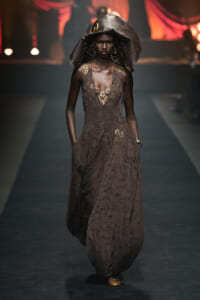 Model walking a runway in a brown lace gown with gold embroidery and a large sculptural hat.