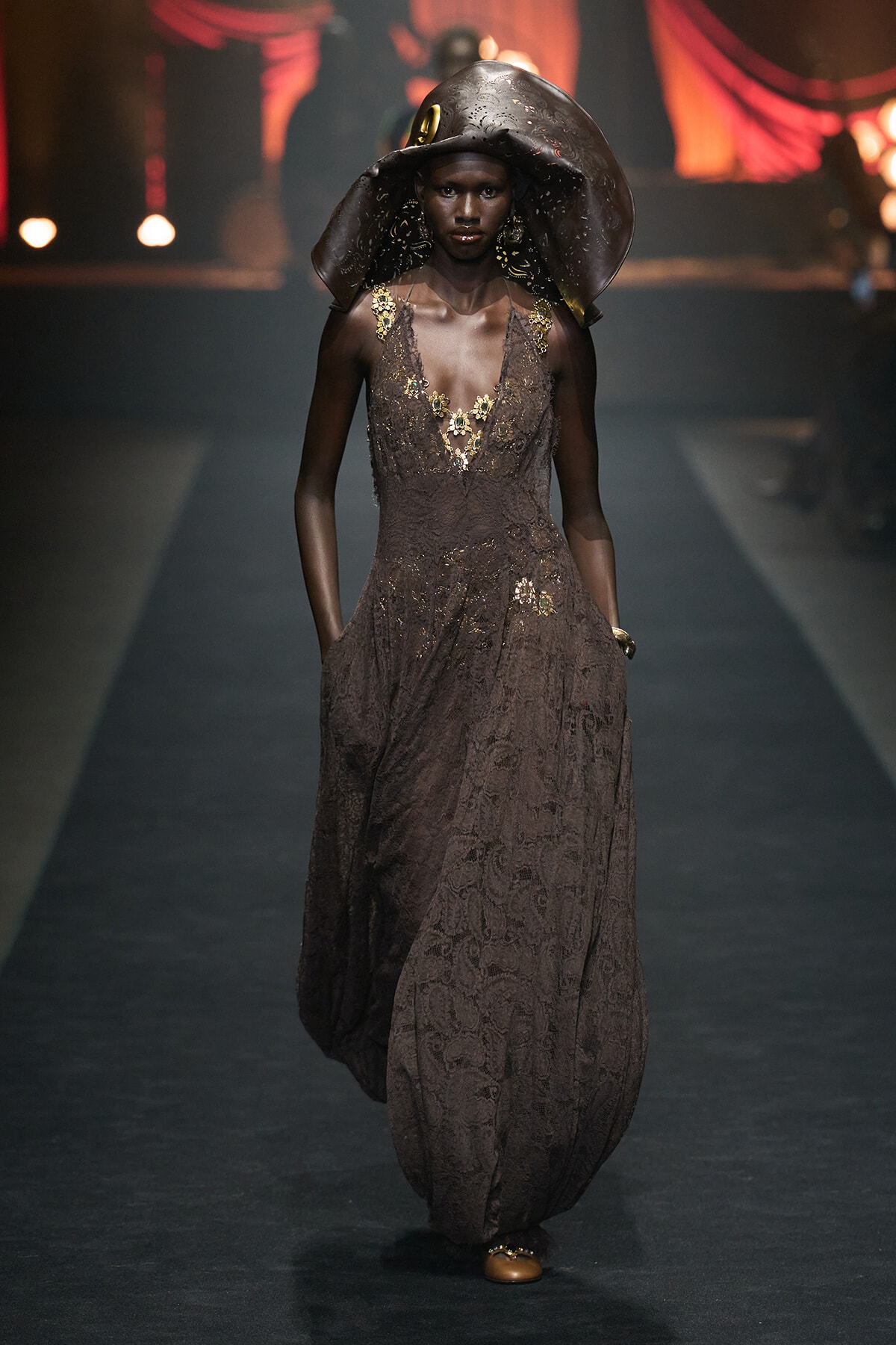 Model walking a runway in a brown lace gown with gold embroidery and a large sculptural hat.