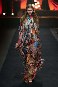 Model walks runway in a long, multicolored fringe gown with metallic tassels and a bead-studded headpiece.