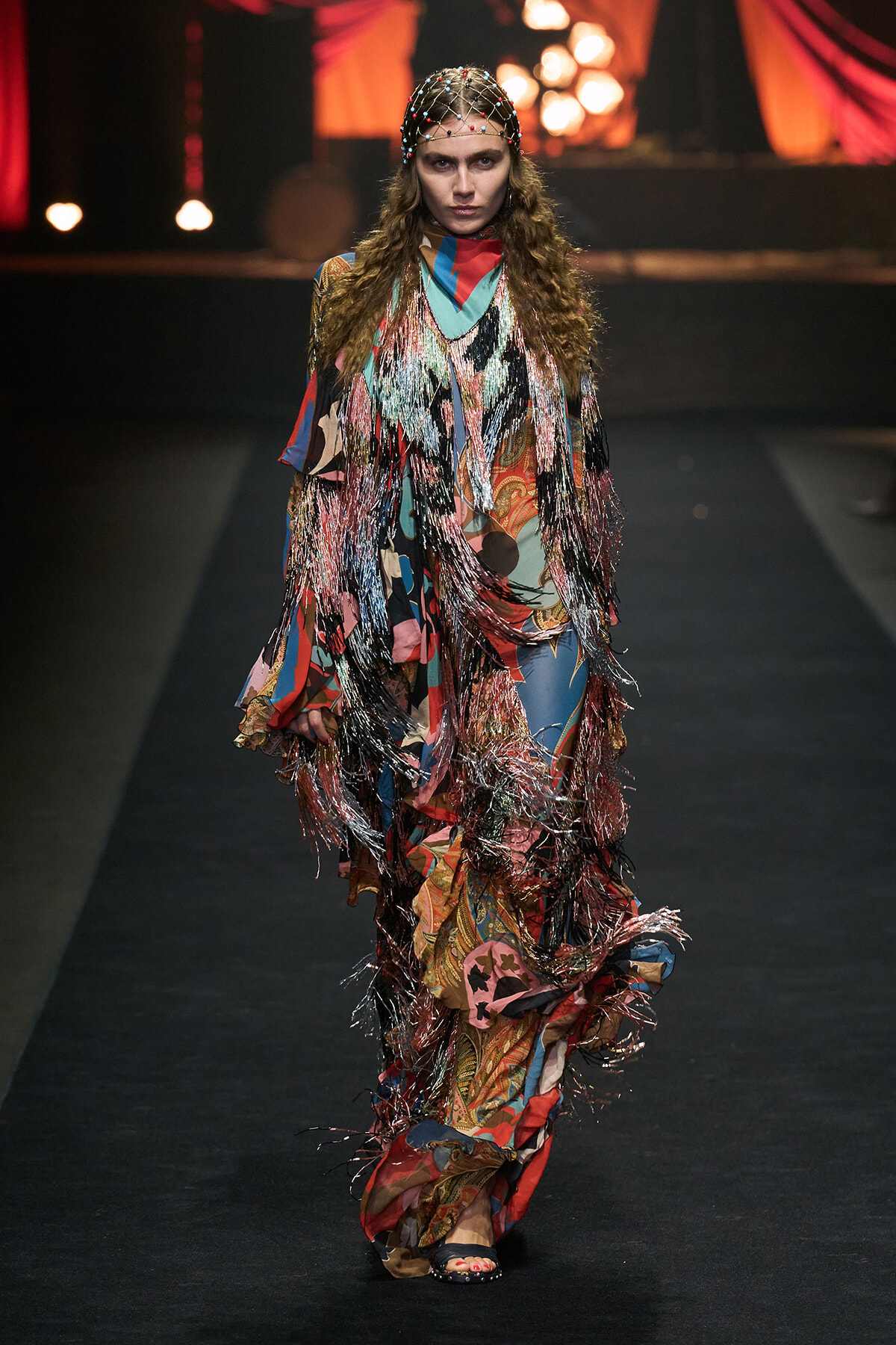 Model walks runway in a long, multicolored fringe gown with metallic tassels and a bead-studded headpiece.