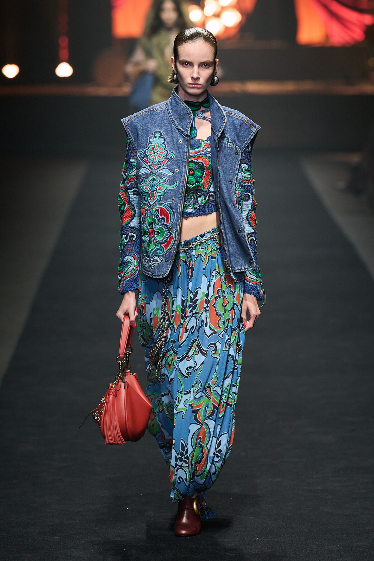 Model walking a fashion show in a blue embroidered denim vest over a colorful paisley outfit, carrying a red handbag.