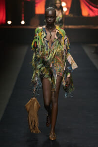 Model walks a runway in a colorful fringe dress with tasseled fringe, carrying a brown fringed bag and wearing layered necklaces and large hoops.