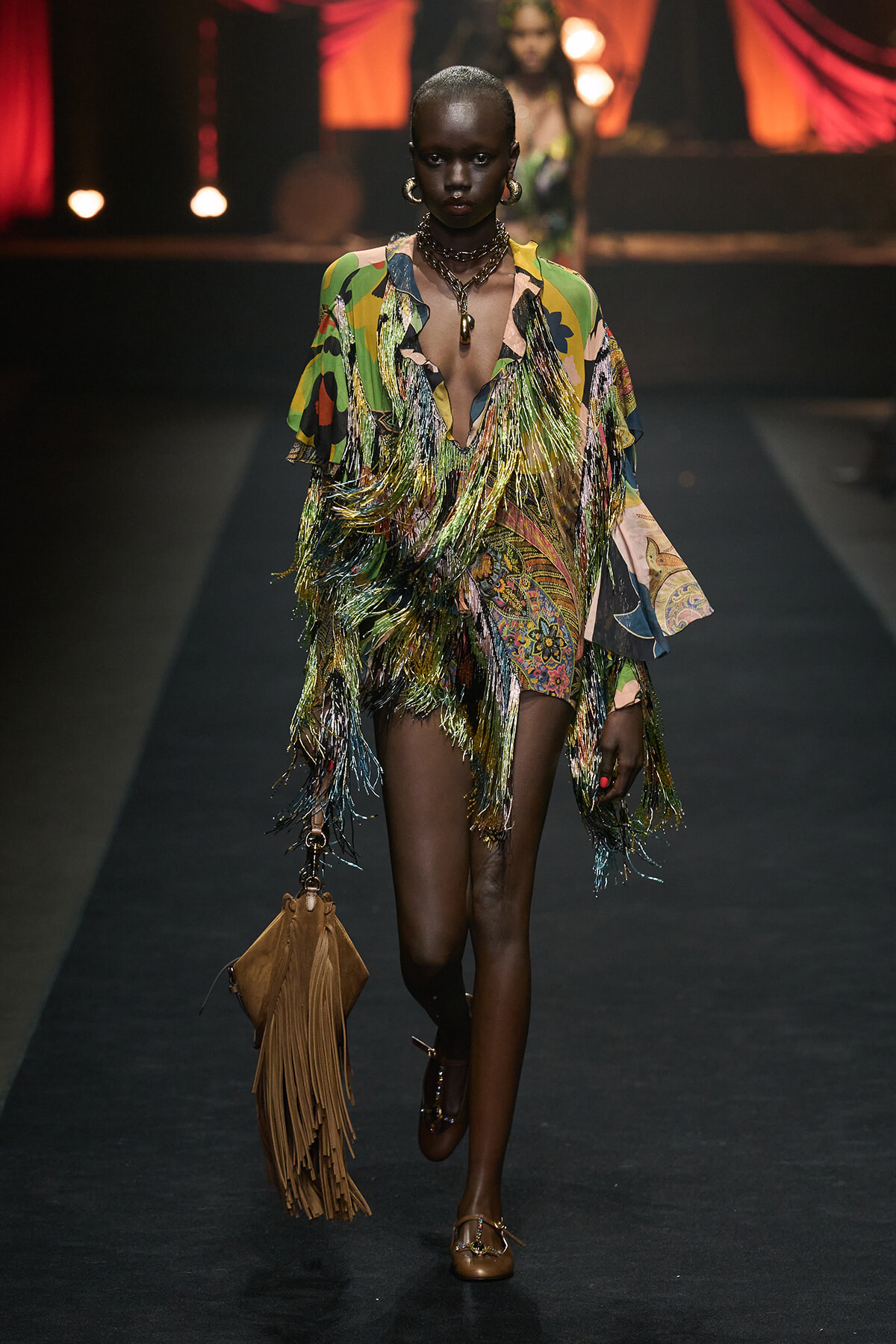 Model walks a runway in a colorful fringe dress with tasseled fringe, carrying a brown fringed bag and wearing layered necklaces and large hoops.