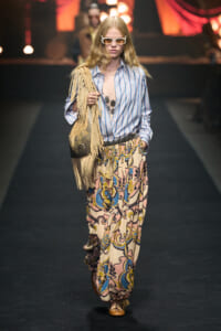 Model walking runway in a boho outfit: striped blue shirt, patterned wide-leg pants, tan fringe bag, oversized white sunglasses.