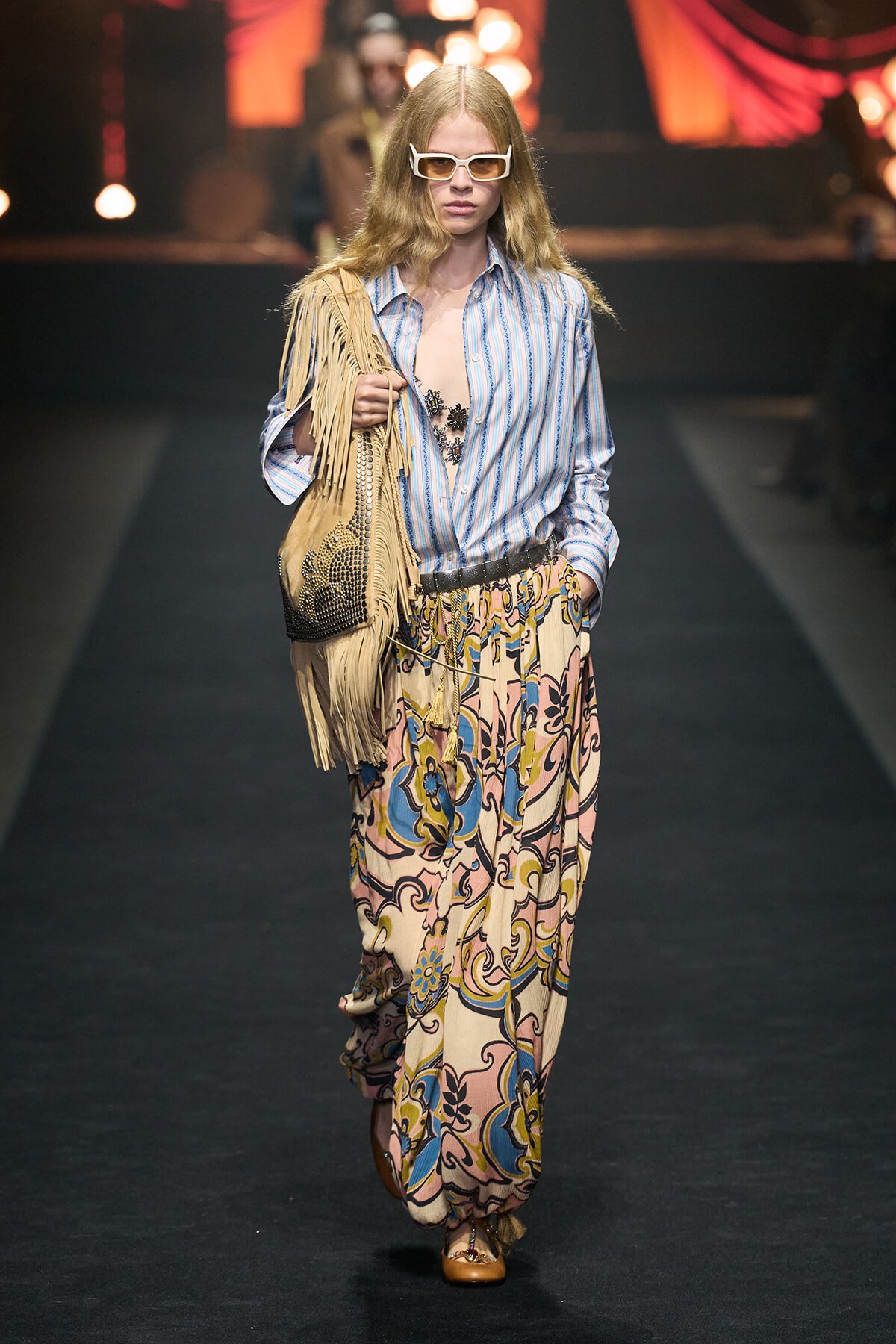 Model walking runway in a boho outfit: striped blue shirt, patterned wide-leg pants, tan fringe bag, oversized white sunglasses.