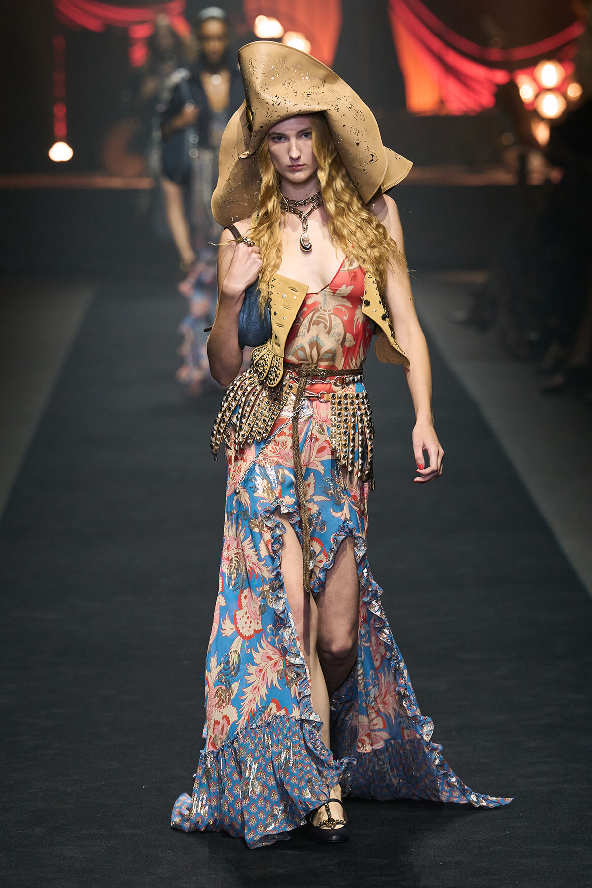 Model walking a fashion runway in a wide-brimmed desert hat, floral maxi dress with a studded belt and ruffled hem.