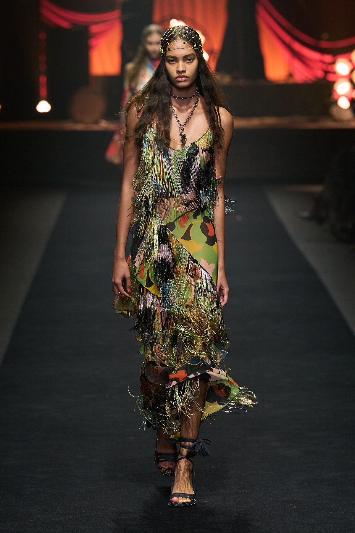 Runway model wearing a colorful, fringed dress with metallic fringe and a beaded headpiece on a dark fashion show runway.
