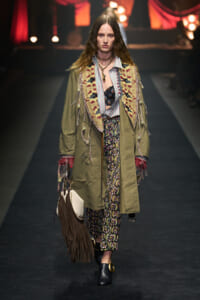 Model walking a runway in an oversized olive coat with a fringed knit vest, patterned pants, and fringe bag. A boho, layered ensemble on display.