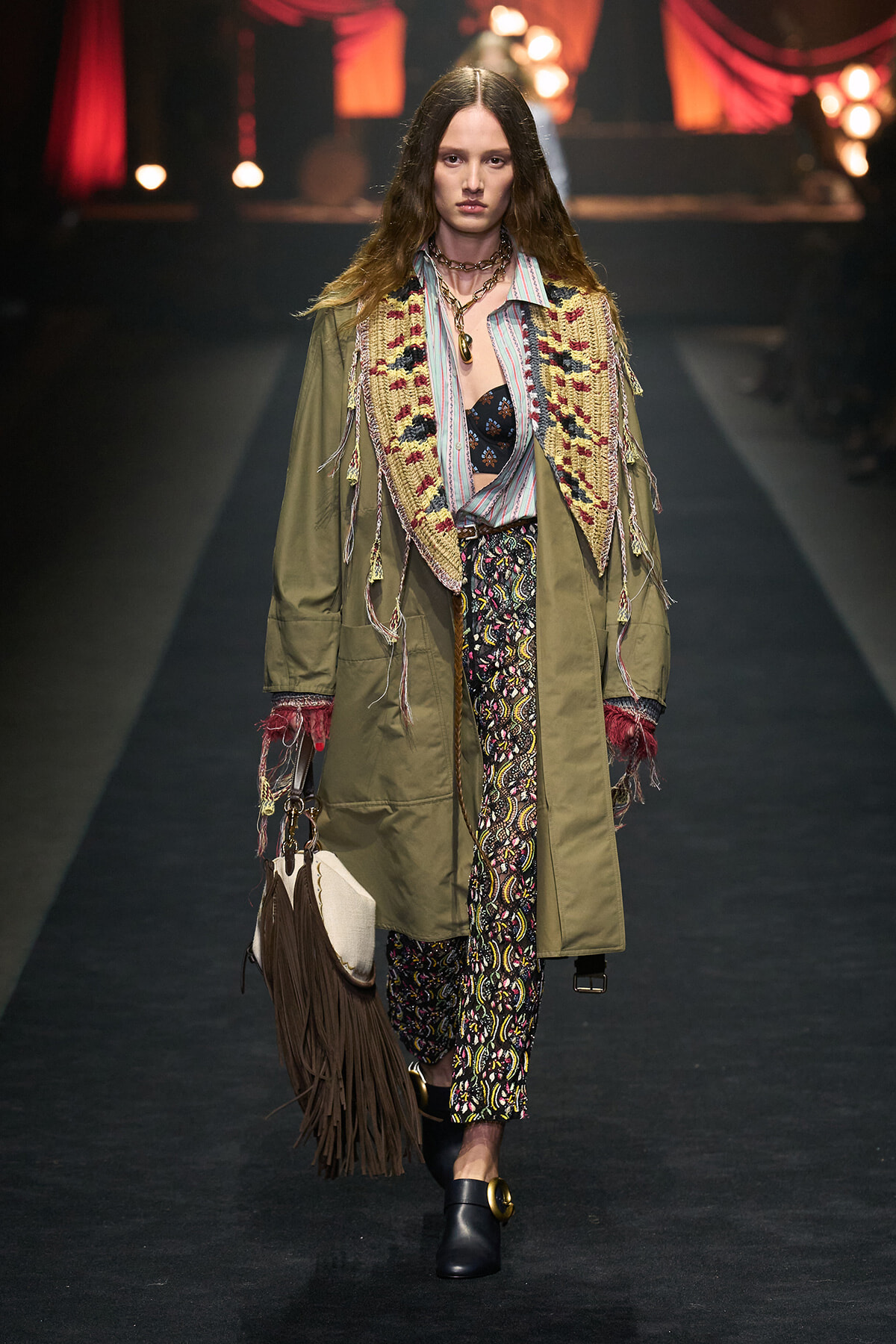 Model walking a runway in an oversized olive coat with a fringed knit vest, patterned pants, and fringe bag. A boho, layered ensemble on display.