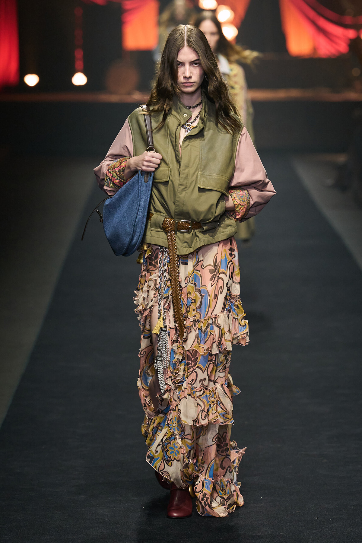 Model walks a fashion runway wearing a layered olive vest over a pink-sleeved blouse and a long, ruffled floral skirt with a braided belt and red boots, carrying a blue handbag.