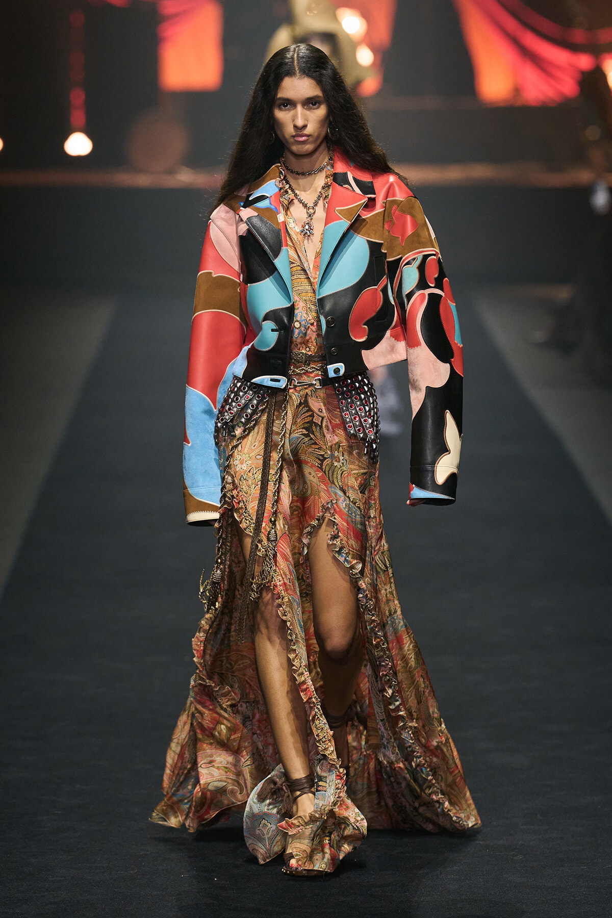 Model walking down a runway in a colorful patchwork jacket over a patterned, high-slit gown with beaded accessories and layered jewelry.