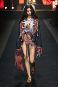 Female model walking a fashion runway in a boho fringe outfit with a blue denim shirt, patterned vest, belt, and fringed bag