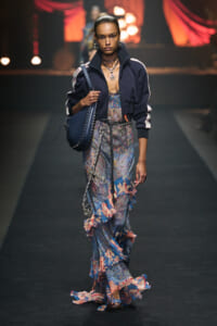Model walking a runway in a navy bomber jacket over a blue paisley maxi dress with a ruffled hem, carrying a navy bag with studded details.