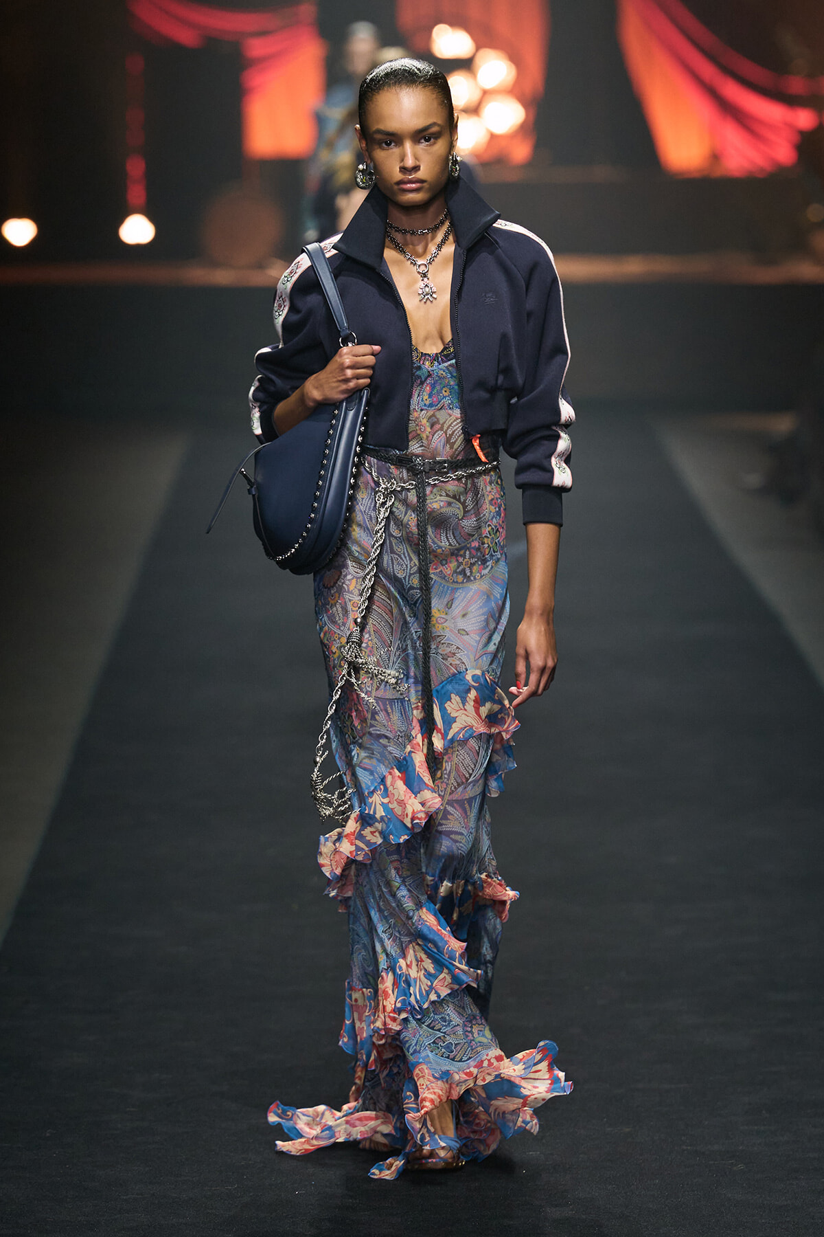 Model walking a runway in a navy bomber jacket over a blue paisley maxi dress with a ruffled hem, carrying a navy bag with studded details.