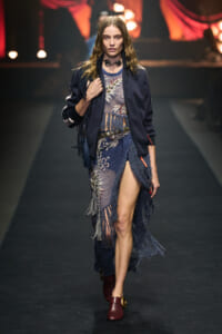 Runway model walking in a navy fringed dress with floral knit details, a dark jacket, and burgundy shoes under stage lights.