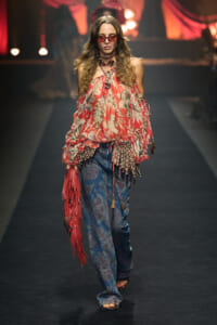 Model walking a fashion runway in a red floral off-shoulder top, blue paisley jeans, bold jewelry, and red sunglasses with a pink fringe bag on her side.