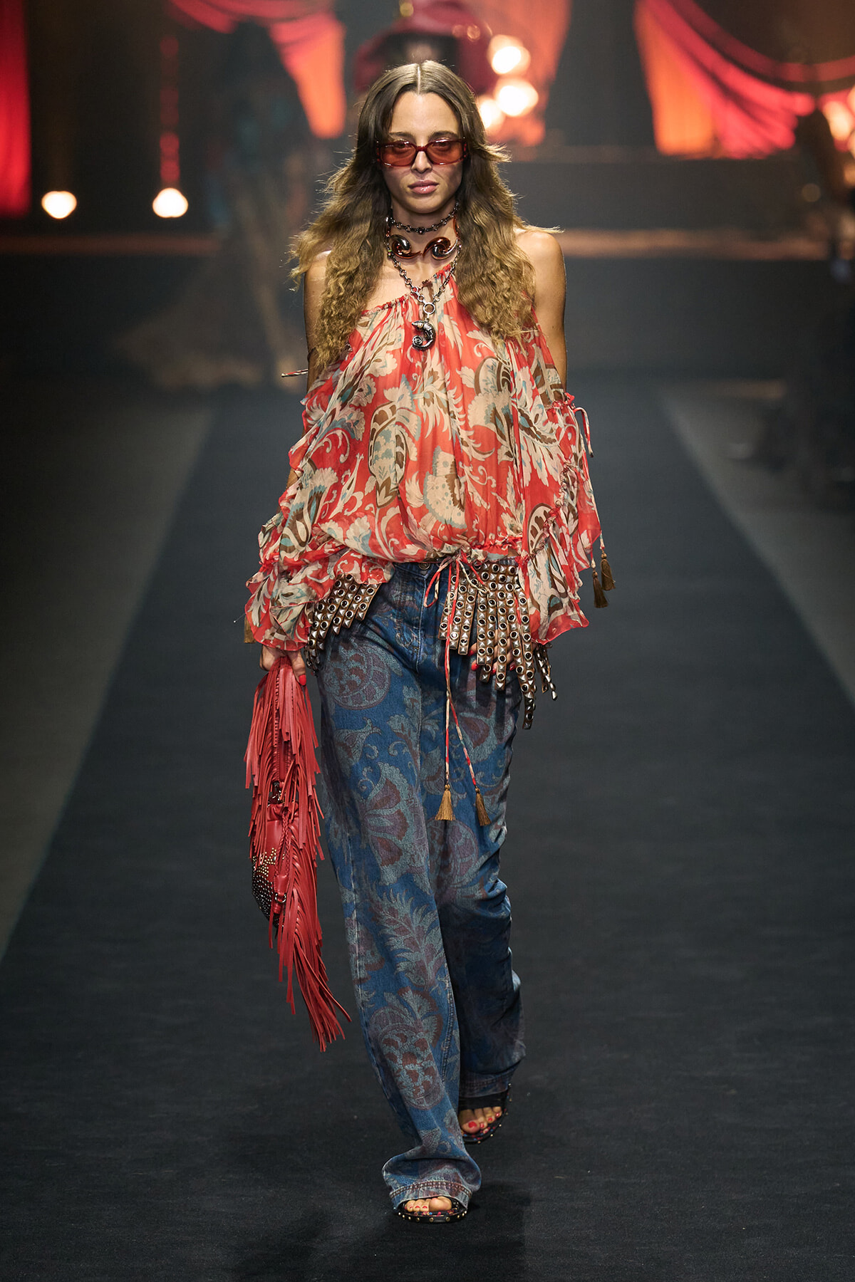 Model walking a fashion runway in a red floral off-shoulder top, blue paisley jeans, bold jewelry, and red sunglasses with a pink fringe bag on her side.
