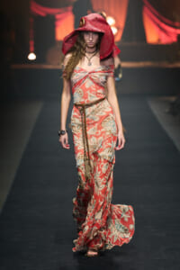 Model walking a fashion show in a red floral off-shoulder gown with a large red hooded hat and braided belt, accessorized with necklaces and bracelets.