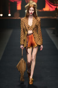 Model walking a runway in a brown patterned blazer with a matching bralette and orange fringe shorts, belt, and a tan fringe handbag down the catwalk.