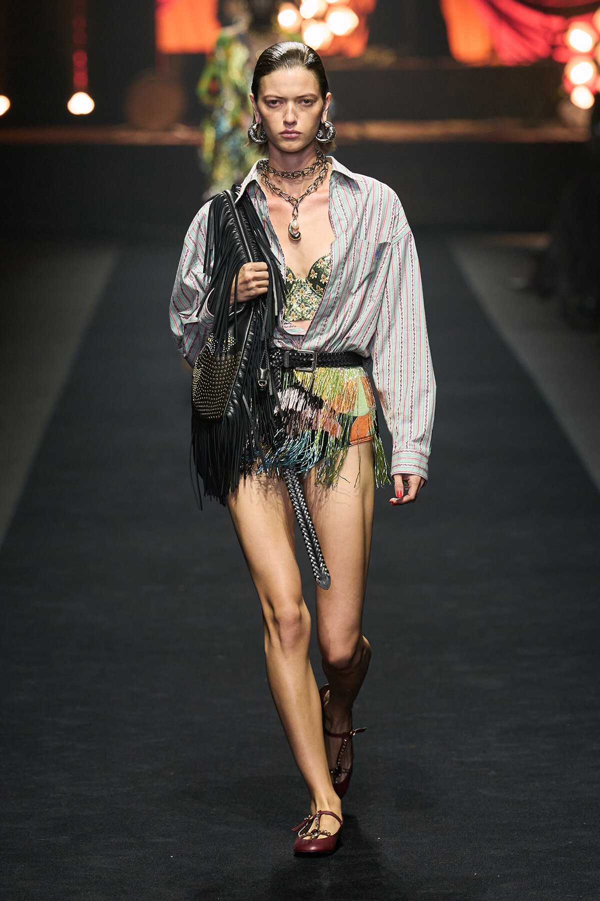 Model walks a fashion runway in a striped oversized shirt, colorful fringed mini-skirt, and bold layered jewelry, carrying a large black fringed bag.