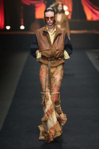 Model walks the runway in a brown and tan leather vest over a yellow striped shirt, paired with a bohemian paisley maxi skirt and belt accessories.