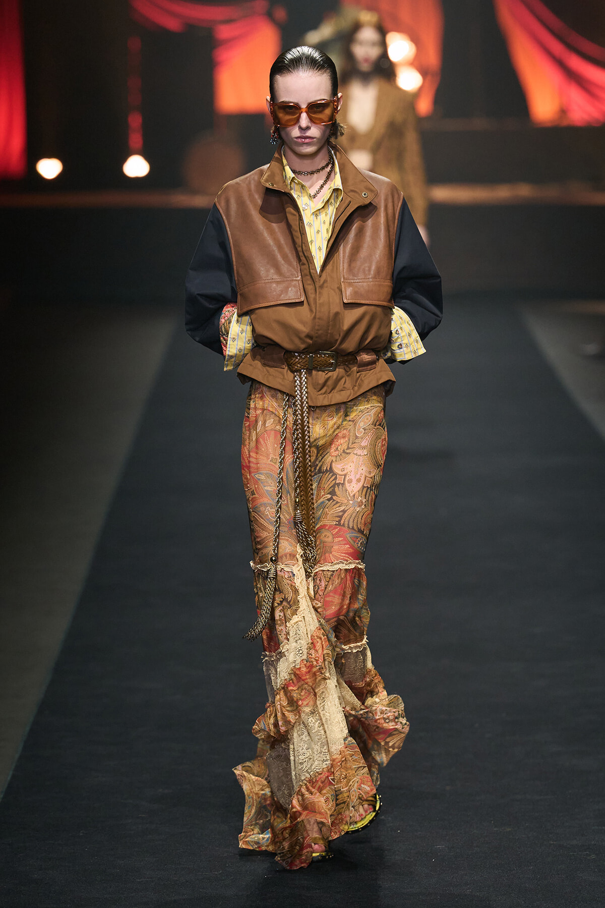 Model walks the runway in a brown and tan leather vest over a yellow striped shirt, paired with a bohemian paisley maxi skirt and belt accessories.